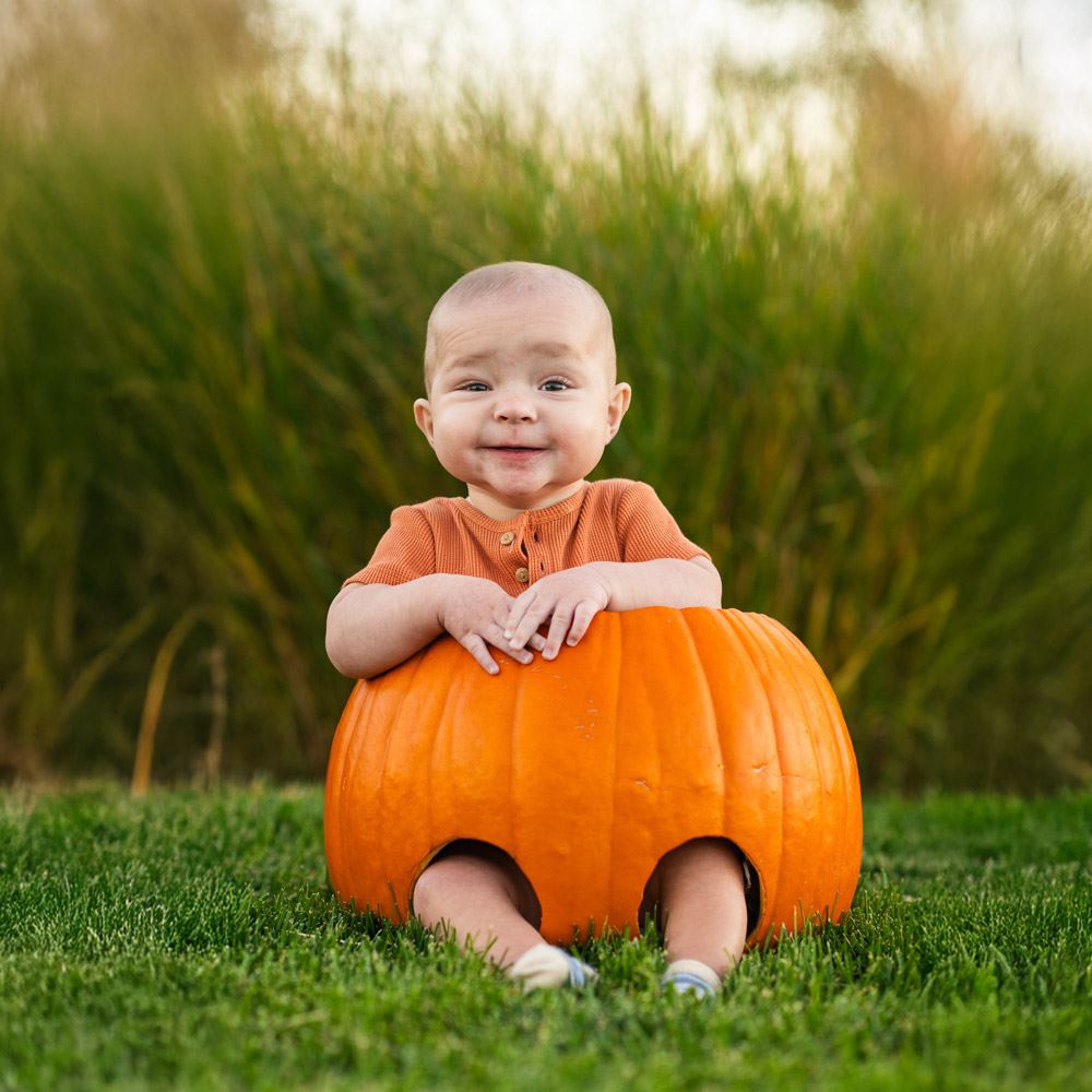 baby wearing an orange shirt sitting inside a pumpkin in a grass area